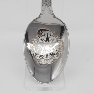 May include: A silver spoon with an engraved pentagram design. The spoon features a detailed floral border around the pentagram. The handle has an ornate, decorative design. The spoon is displayed on a clear stand against a white background.