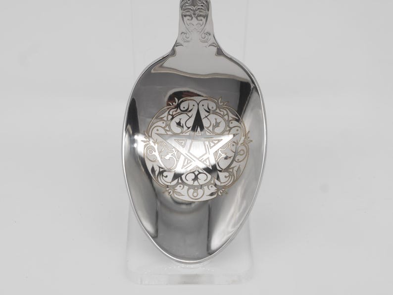 May include: A silver spoon with an engraved pentagram design. The spoon features a detailed floral border around the pentagram. The handle has an ornate, decorative design. The spoon is displayed on a clear stand against a white background.