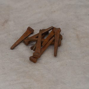 May include: A collection of rusty, antique cut nails. These nails have a rectangular head and a tapered, square shank. The nails are scattered on a neutral-toned, textured surface, showcasing their aged, weathered appearance. Ideal for rustic decor or restoration projects.