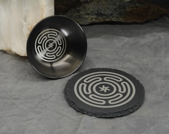Könnte beinhalten: Eine schwarze Metallschale und ein passender runder Untersetzer aus schwarzem Schiefer, beide mit einem silberfarbenen, eingravierten Hekate-Rad-Symbol. Die Schale hat eine glatte, reflektierende Innenfläche, während der Untersetzer einen strukturierten Rand aufweist.