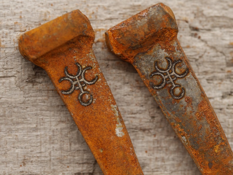 May include: Two weathered, rusty metal objects with a rectangular shape and a decorative silver design. The objects have a worn, orange-brown color, suggesting age and exposure to the elements. The design is a stylized, symmetrical pattern.
