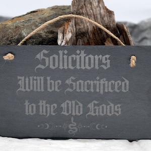 May include: A rectangular, dark gray slate sign with the text "Solicitors Will be Sacrificed to the Old Gods" in a gothic font. The sign has a jute rope for hanging and is displayed against a natural background.