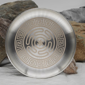 May include: A silver metal plate with a decorative design of a Greek key border and a concentric circle pattern in the center. The design is in a light gold color.