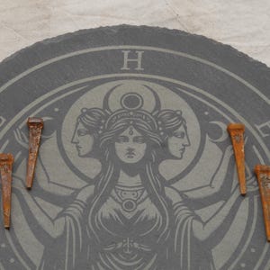 May include: A dark gray, circular slate with a detailed etched design featuring a central female figure with three faces, surrounded by celestial symbols. Five rusty, aged nails are arranged around the design.