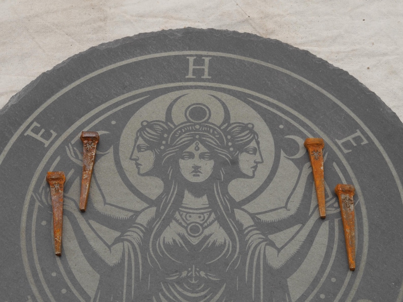 May include: A dark gray, circular slate with a detailed etched design featuring a central female figure with three faces, surrounded by celestial symbols. Five rusty, aged nails are arranged around the design.
