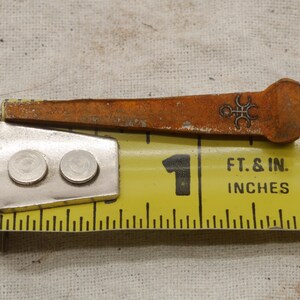 May include: Close-up of a rusty, antique nail with a decorative symbol near the head, resting on a yellow measuring tape. The tape shows measurements in inches, with the nail measuring approximately 1 inch in length.