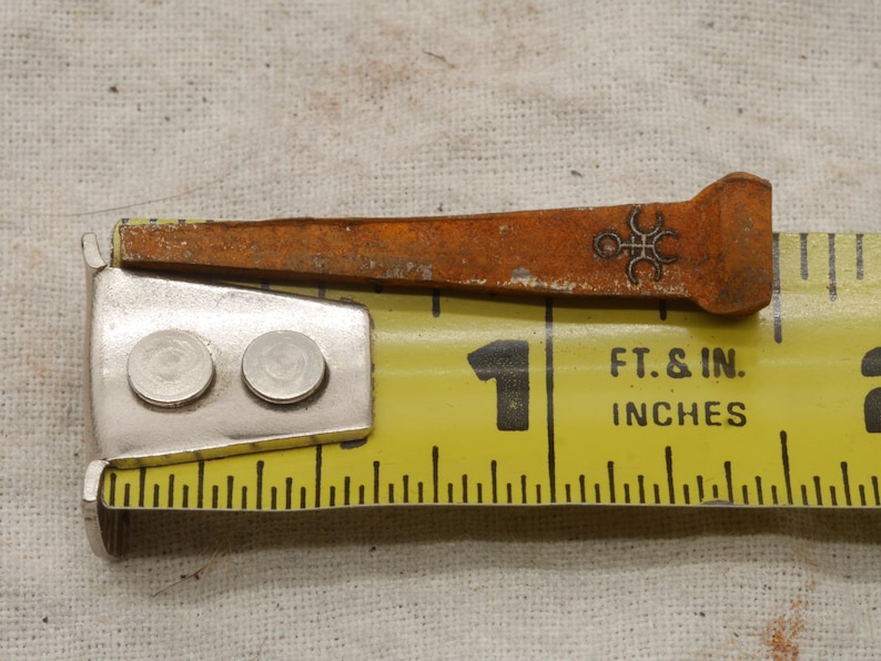 May include: Close-up of a rusty, antique nail with a decorative symbol near the head, resting on a yellow measuring tape. The tape shows measurements in inches, with the nail measuring approximately 1 inch in length.