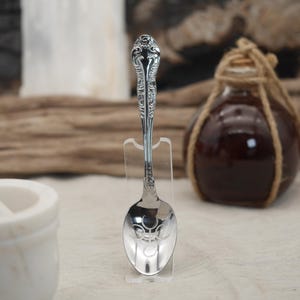 K&ouml;nnte beinhalten: Ein silberner L&ouml;ffel mit Zierdetails an Griff und Schale, pr&auml;sentiert auf einem transparenten St&auml;nder. Der L&ouml;ffel ist hochglanzpoliert. Im Hintergrund eine dunkle Glasflasche mit Kordel.