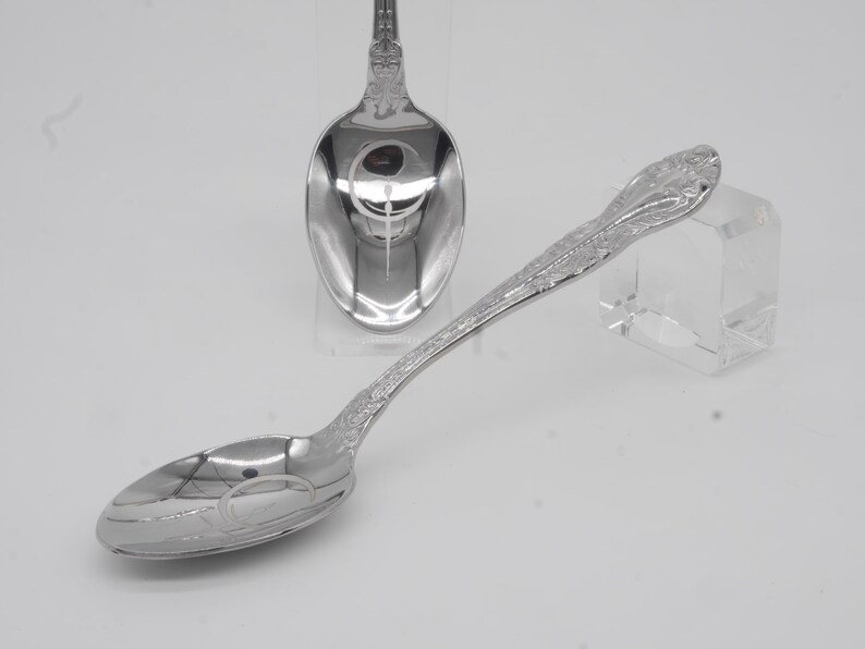 May include: Two ornate silver spoons with a floral design on the handles and oval bowls. The spoons are highly polished, reflecting the light. One spoon is in the foreground, the other in the background.