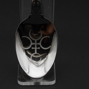K&ouml;nnte beinhalten: Ein silberner L&ouml;ffel mit einem verzierten Griff und einer polierten Laffe. Die Laffe weist ein ge&auml;tztes Design eines Symbols mit Halbmondformen und einem kreuzartigen Element auf. Der L&ouml;ffel wird vor einem dunklen Hintergrund pr&auml;sentiert.