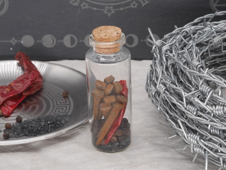 May include: A clear glass bottle with a cork stopper, filled with brown objects, red peppers, and black peppercorns. A silver plate with red peppers and black salt is to the left. A coil of barbed wire is on the right.
