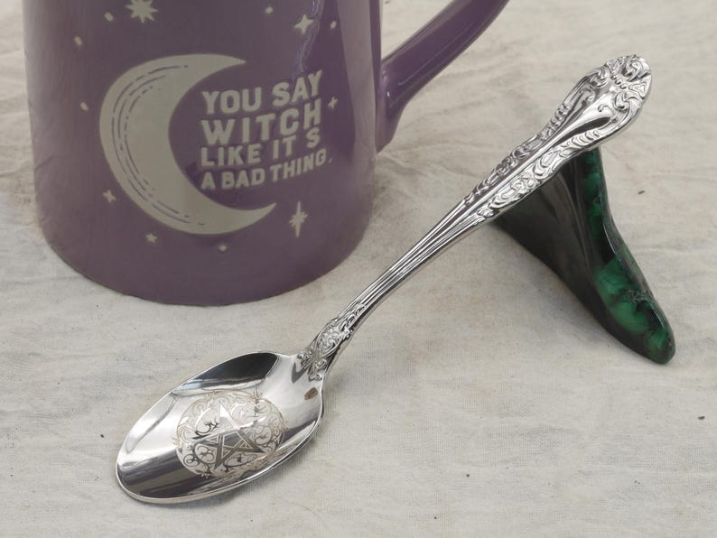 May include: A silver spoon with an engraved pentagram design on the bowl, resting on a green stand. A purple mug with the text "YOU SAY WITCH LIKE IT'S A BAD THING" is in the background. The spoon has ornate detailing.