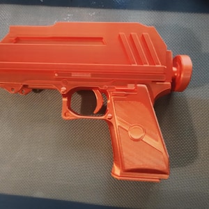 May include: An orange 3D-printed toy gun with a detailed design. The toy gun features a trigger, grip, and a cylindrical component at the end. The object is placed on a gray surface.