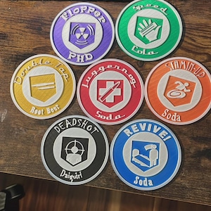 Peut inclure: Ensemble de sept dessous de verre ronds et colorés, chacun avec un design et une couleur distincts. Les dessous de verre présentent des graphiques et du texte tels que "PHD", "Speed Cola" et "Double Tap Root Beer". Ils sont disposés sur une surface en bois.