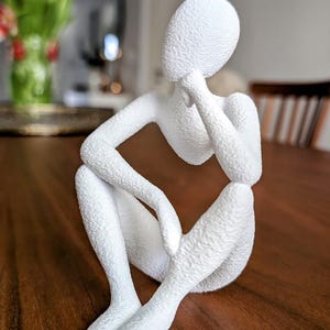 Könnte beinhalten: Weiße abstrakte Skulptur einer sitzenden Person, die das Kinn auf die Hand legt. Die Skulptur hat eine strukturierte Oberfläche und ist aus glattem Material gefertigt.