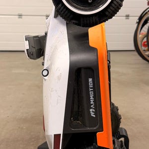 May include: A white and orange robotic lawn mower with black wheels featuring small treaded rollers, positioned vertically on a black plastic charging station. The side of the unit displays the brand name MAMMOTION in white text.