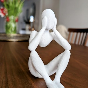 Könnte beinhalten: Weiße abstrakte Skulptur einer Person, die mit dem Kopf in den Händen sitzt. Die Skulptur ist aus einem glatten, strukturierten Material gefertigt und steht auf einer Holzoberfläche.