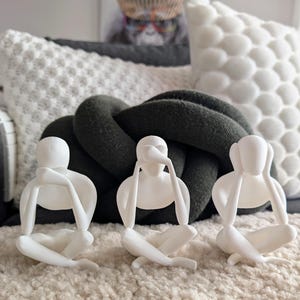 May include: Three white figurines of people covering their eyes, ears, and mouth. The figurines are sitting on a white fluffy surface.