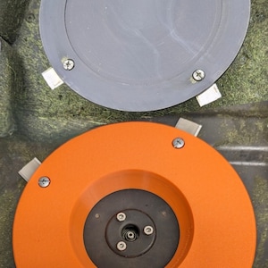 May include: Two round, flat, metal mower blades. The top blade is grey and the bottom blade is orange. Both blades have a centre hole and multiple screw holes around the edge.