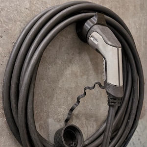 Może przedstawiać: Czarny kabel do ładowania pojazdów elektrycznych z szarym złączem z napisem "Schneider Electric". Kabel jest zwinięty i gotowy do użycia.