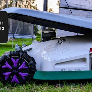 May include: A gray robotic lawn mower shelter covering a white robotic mower with green accents and purple wheel covers. A black sign on the left lists compatibility: COMPATIBLE WITH: LUBA 1, LUBA 2, LUBA 2X, LUBA 3, LUBA MINI 1, and LUBA MINI 2.