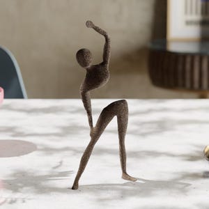 Könnte beinhalten: Eine braune abstrakte Skulptur einer Person in einer Tanzpose. Die Skulptur ist aus einem rauen, strukturierten Material gefertigt und steht auf einer weißen Oberfläche.