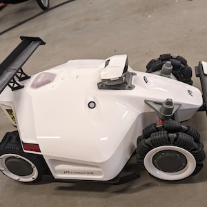 Pode incluir: Um cortador de relva robótico branco com um spoiler preto e grandes rodas pretas. O cortador de relva tem um pequeno sensor branco no topo e o texto "E.S. AMMOWORX" no lado.