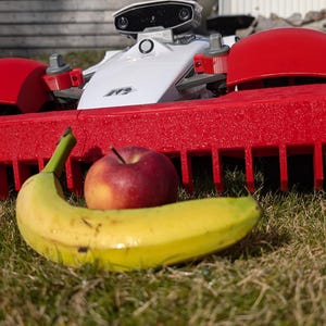 May include: A red and white robotic lawnmower with a red plastic blade guard. A yellow banana and a red apple are in front of the lawnmower on a green grassy surface.