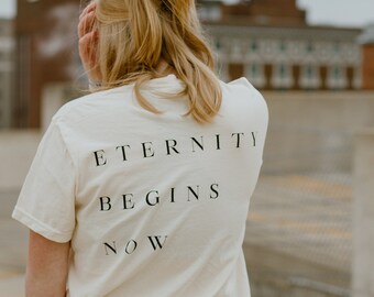 May include: A person wearing a white t-shirt with the text "Eternity Begins Now" printed on the back. The person is wearing blue jeans and has their hair pulled back in a ponytail.