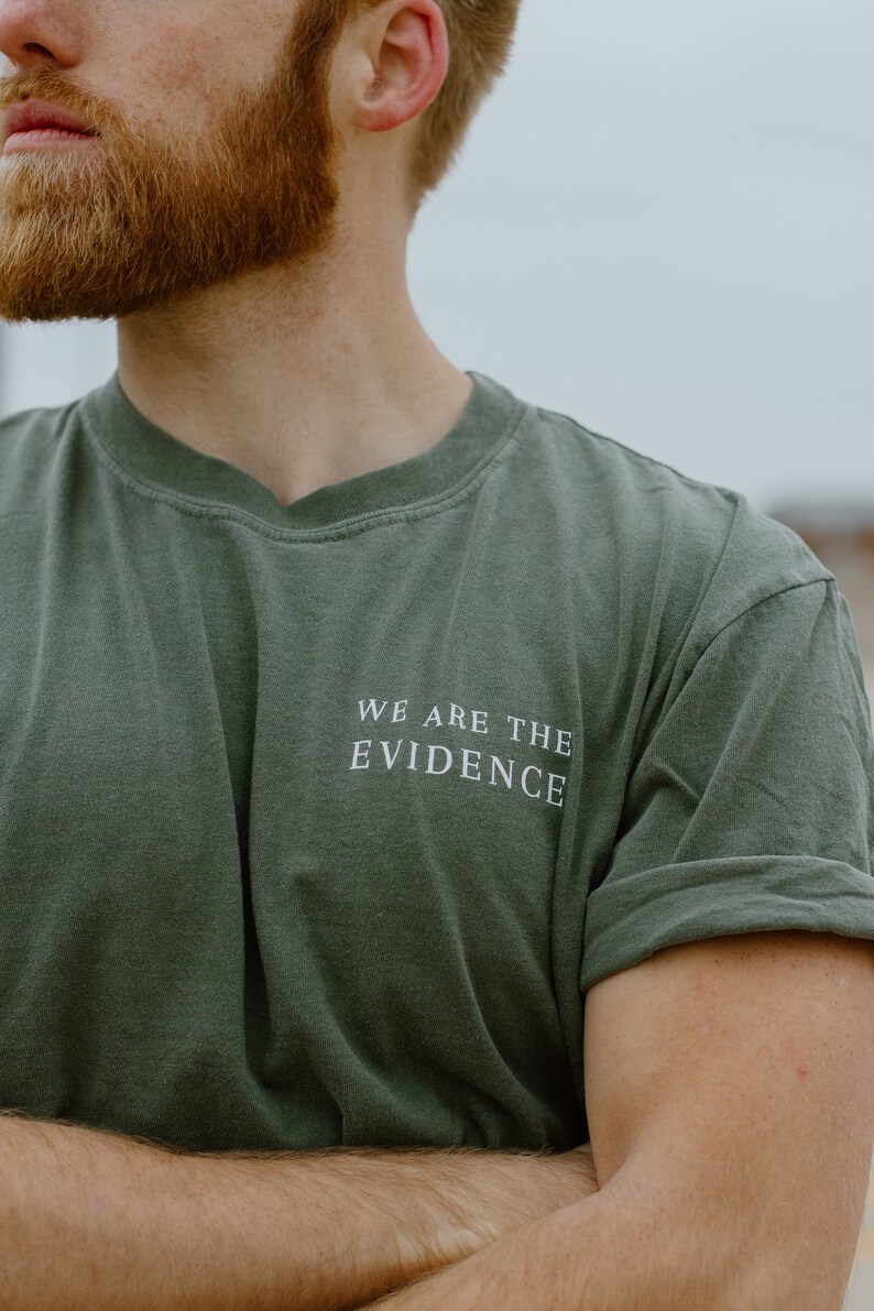 May include: Olive green t-shirt with the text "WE ARE THE EVIDENCE" in white. The shirt has a crew neck and short sleeves, with the sleeves rolled up. The image is a close-up of the shirt and upper body.