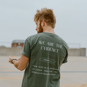 May include: Olive green t-shirt with the text "WE ARE THE EVIDENCE" and a Bible verse. The shirt has rolled-up sleeves and a relaxed fit. The text "HEBREWS 11:1" is also visible.
