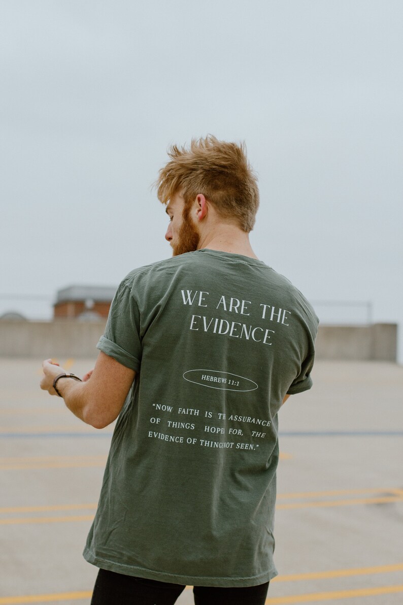 May include: Olive green t-shirt with the text "WE ARE THE EVIDENCE" and a Bible verse. The shirt has rolled-up sleeves and a relaxed fit. The text "HEBREWS 11:1" is also visible.