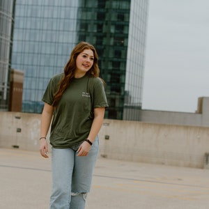 May include: An olive green t-shirt with the text "WE ARE EVIDENCE" is paired with light blue jeans with a distressed knee. The outfit is completed with white sneakers. The background features a modern glass building.