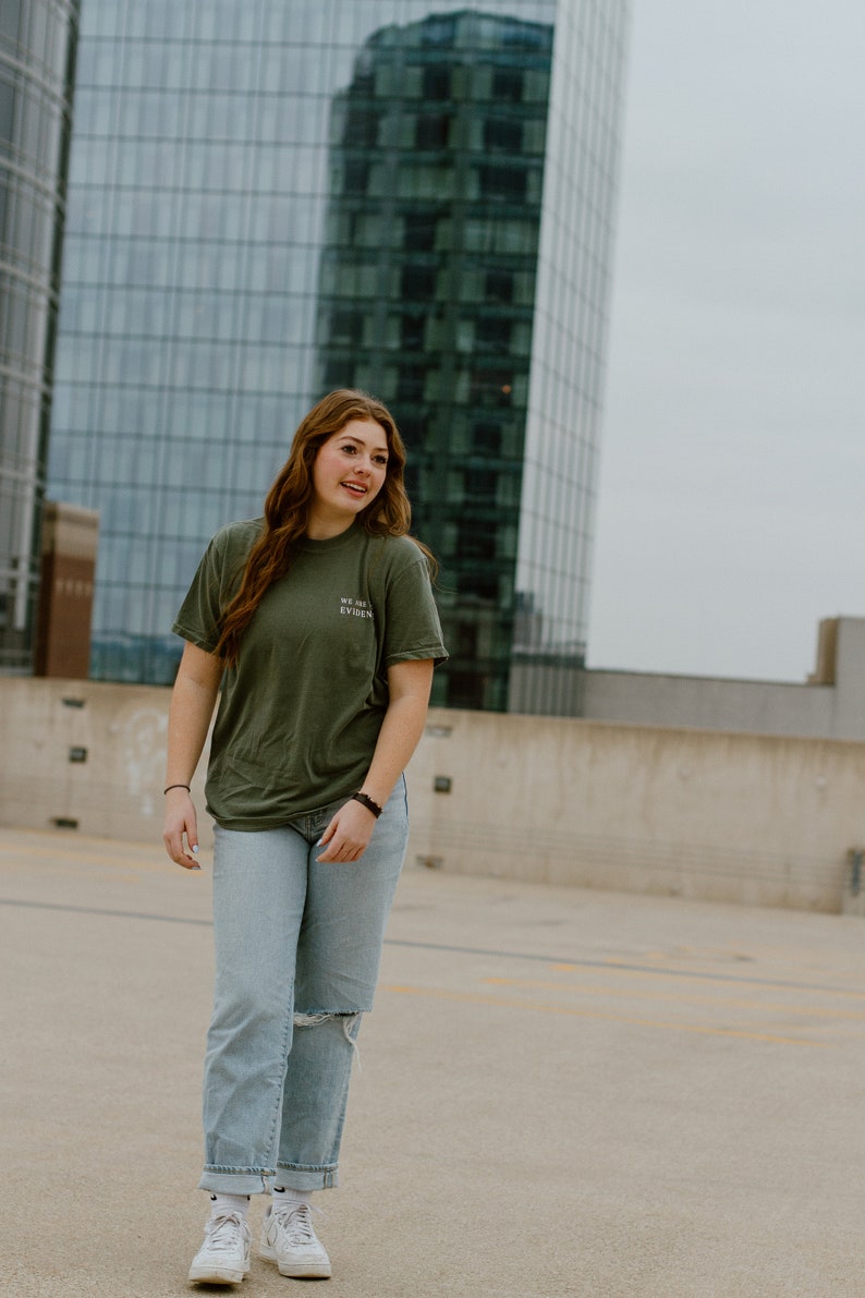 May include: An olive green t-shirt with the text "WE ARE EVIDENCE" is paired with light blue jeans with a distressed knee. The outfit is completed with white sneakers. The background features a modern glass building.