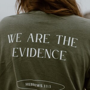 May include: Olive green t-shirt with the text "WE ARE THE EVIDENCE" in white, with "HEBREWS 11:1" below. The shirt is a solid color and has a classic crew neck.