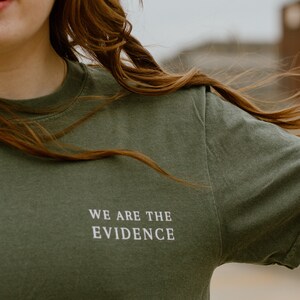 May include: Olive green t-shirt with the text "WE ARE THE EVIDENCE" in white. The shirt is a crew neck style and appears to be made of a soft material. The image shows the upper body of a person wearing the shirt.