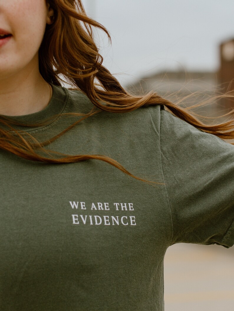 May include: Olive green t-shirt with the text "WE ARE THE EVIDENCE" in white. The shirt is a crew neck style and appears to be made of a soft material. The image shows the upper body of a person wearing the shirt.