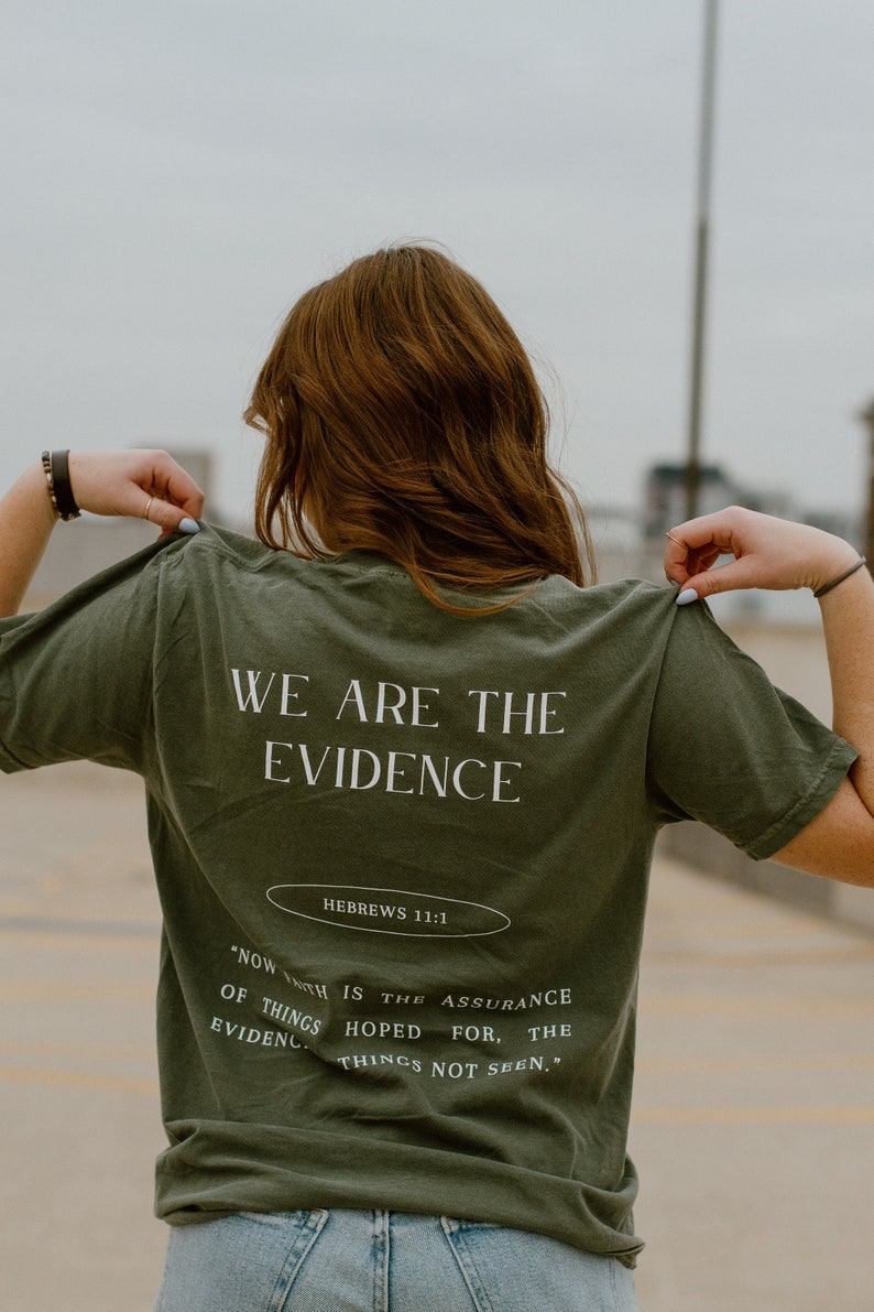 May include: Olive green t-shirt with the text "WE ARE THE EVIDENCE" and "HEBREWS 11:1" printed in white. The shirt also features a quote about faith. The person is wearing the shirt.