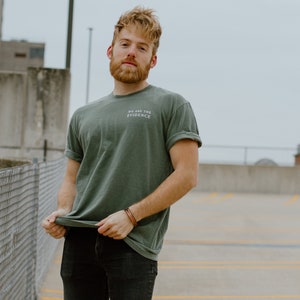 May include: A person wearing a green t-shirt with the text "WE ARE THE EVIDENCE" in white. The person is also wearing black jeans and a brown leather bracelet. The background is a parking garage.