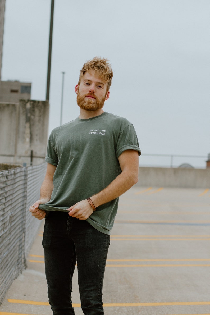 May include: A person wearing a green t-shirt with the text "WE ARE THE EVIDENCE" in white. The person is also wearing black jeans and a brown leather bracelet. The background is a parking garage.