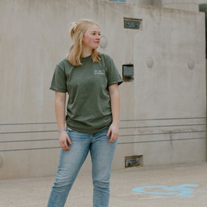 May include: An olive green t-shirt with the text "WE ARE THE EVIDENCE" is paired with light blue jeans and white high-top sneakers. The person is standing in front of a concrete building.
