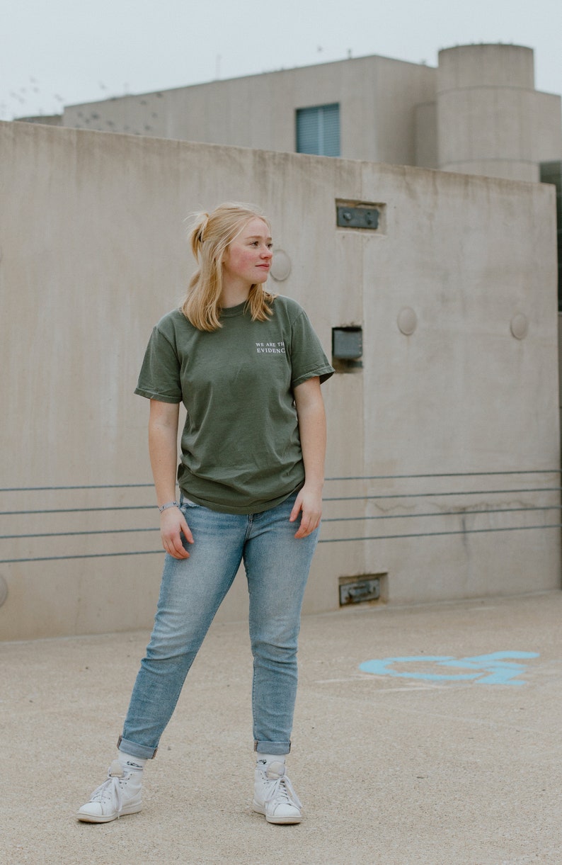 May include: An olive green t-shirt with the text "WE ARE THE EVIDENCE" is paired with light blue jeans and white high-top sneakers. The person is standing in front of a concrete building.