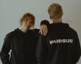 May include: Two people wearing black hooded sweatshirts. The person in the back has the word "PURSUE" printed in white on the back of their sweatshirt.