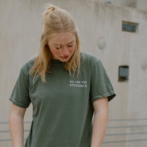 May include: An olive green t-shirt with the text "WE ARE THE EVIDENCE" in white. The shirt is worn by a person wearing blue jeans. The background is a neutral-toned concrete wall.