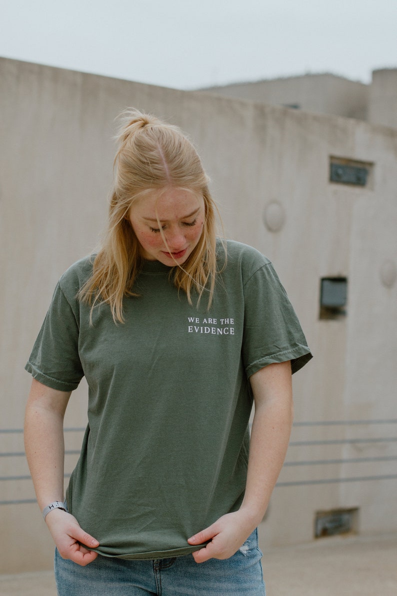 May include: An olive green t-shirt with the text "WE ARE THE EVIDENCE" in white. The shirt is worn by a person wearing blue jeans. The background is a neutral-toned concrete wall.