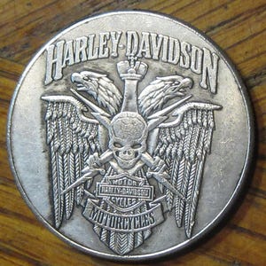 Könnte beinhalten: Silberfarbene Harley Davidson Motorradmünze mit einem Totenkopf mit gekreuzten Blitzen und zwei Adlern mit ausgebreiteten Flügeln. Die Münze trägt auch den Text "MOTOR HARLEY-DAVIDSON CYCLES MOTORCYCLES" um den Totenkopf herum.