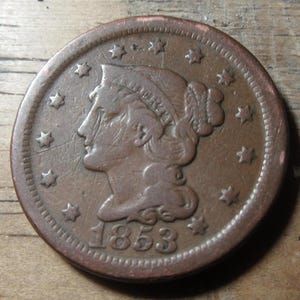 May include: A copper one-cent coin from 1853 with a portrait of Liberty on the obverse. The coin has a ring of stars around the edge.