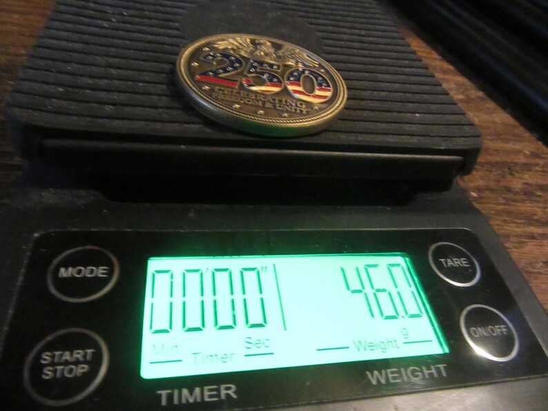 K&ouml;nnte beinhalten: Eine Gedenkm&uuml;nze mit roten, wei&szlig;en und blauen Akzenten, die mit "250 CELEBRATING FREEDOM & UNITY" gekennzeichnet ist, liegt auf einer Digitalwaage. Die Waage zeigt ein Gewicht von 46,0 Gramm an. Die Waage hat eine Timer-Funktion.