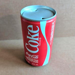 May include: A vintage Coca-Cola can in red with a light blue wave design. The word "Coke" is in white script. The can has a silver top and contains 354 ml.