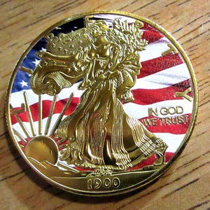 Könnte beinhalten: Eine vergoldete amerikanische Silberdollar-Münze mit einer Darstellung von Lady Liberty, die nach vorne geht und eine Fackel und einen Zweig mit Olivenblättern hält. Die Münze zeigt im Hintergrund ein amerikanisches Flaggen-Design und die Inschrift "In God We Trust". Das Jahr "1900" ist am unteren Rand der Münze eingraviert.